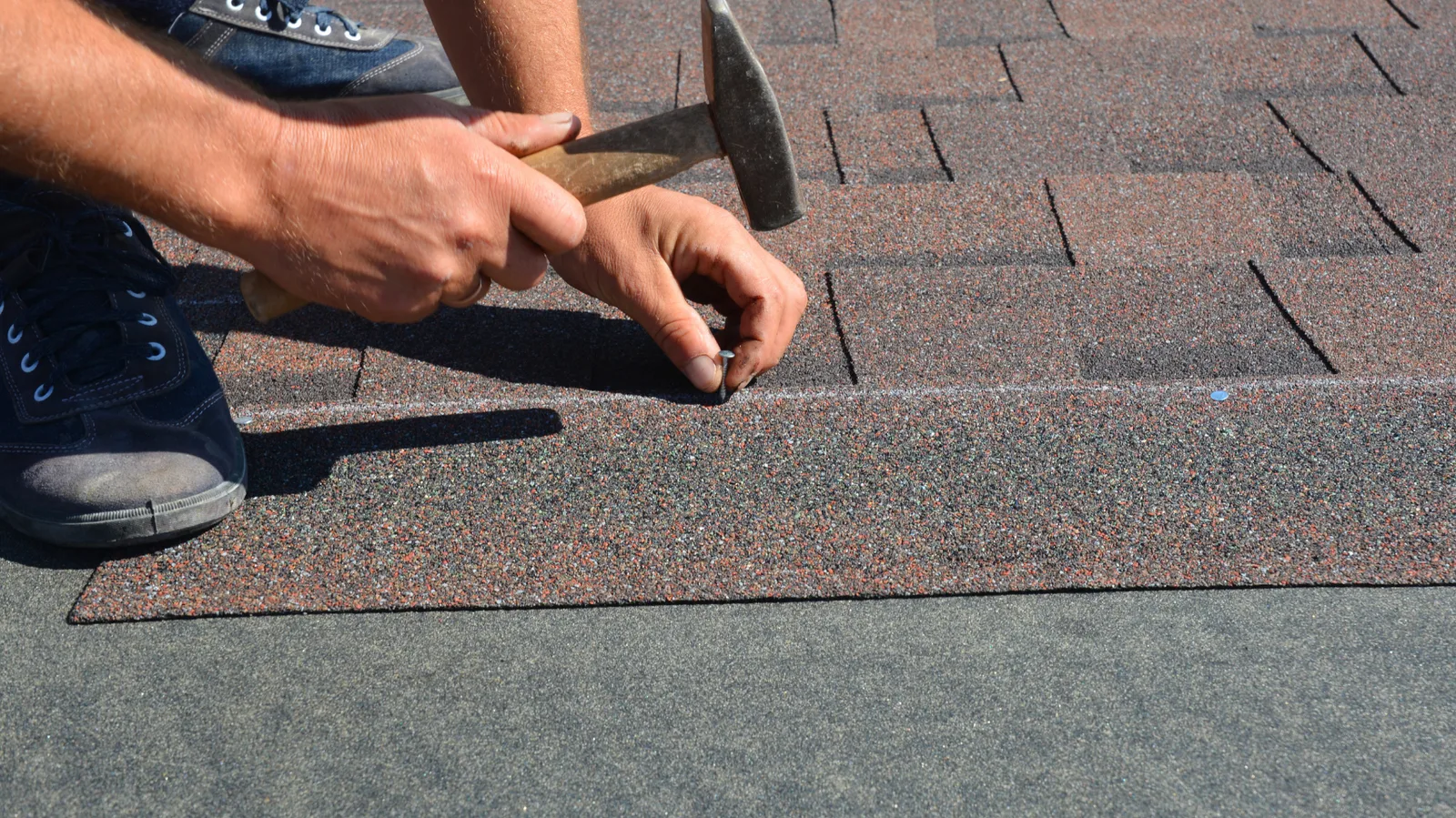 A roofer hammering a nail into an asphalt shingle